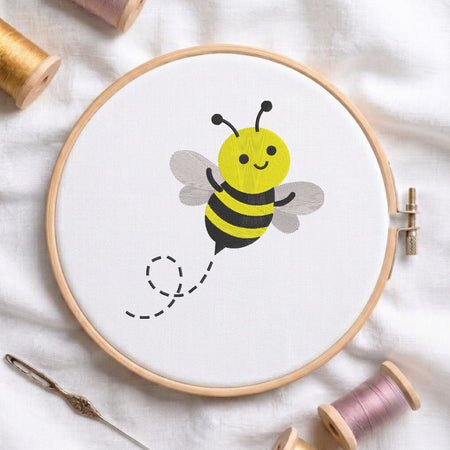 A wooden embroidery hoop holding white fabric with a cute yellow and black bumblebee design, surrounded by spools of thread and a needle on a white fabric background.