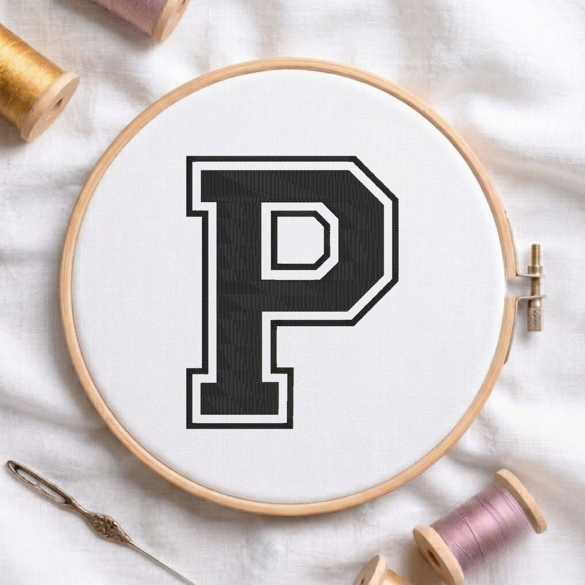 A wooden embroidery hoop holding white fabric with a large black letter P outlined in white. A metal needle lies at the lower left, and spools of thread are arranged around the hoop on a white fabric background.
