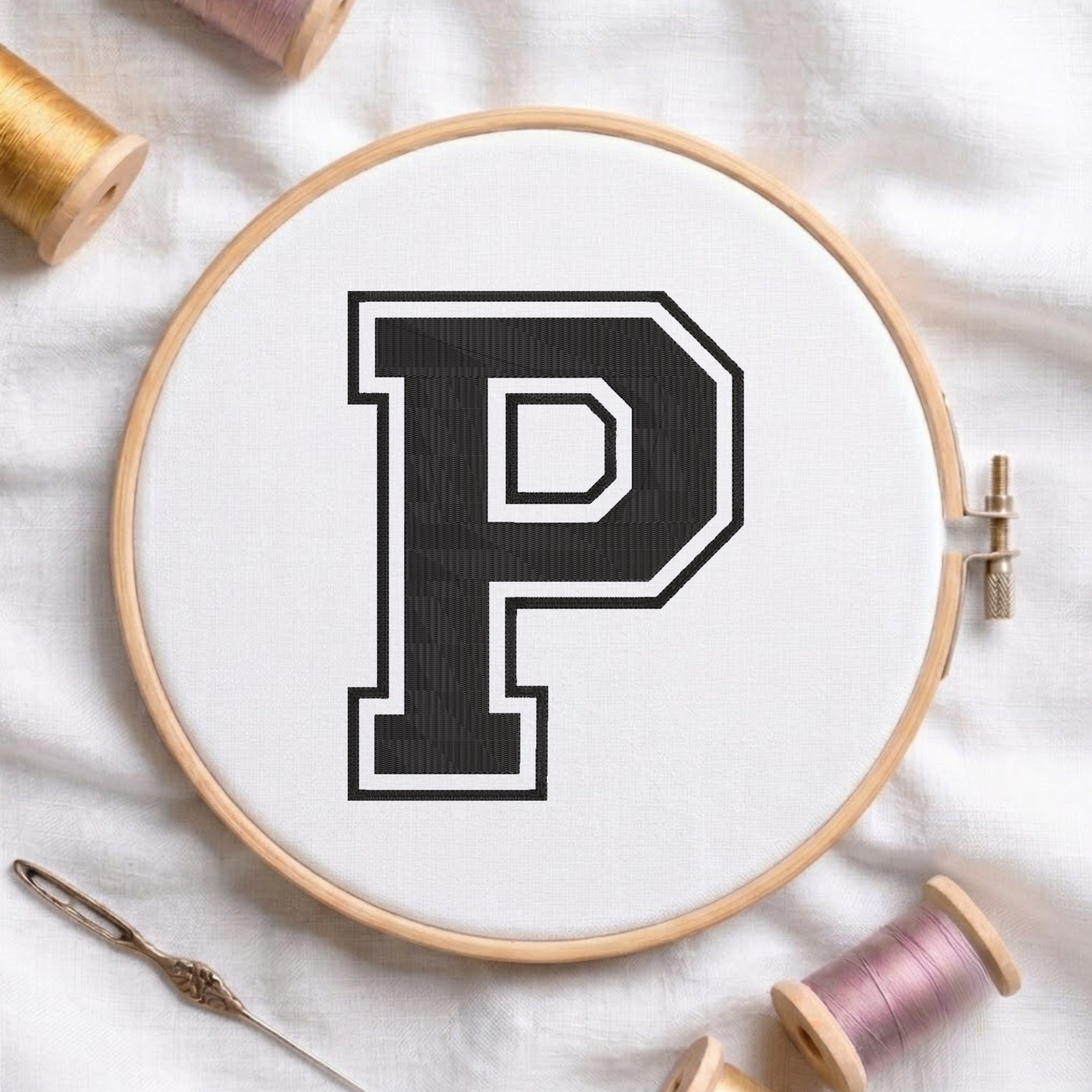 A wooden embroidery hoop holding white fabric with a large black letter P outlined in white. A metal needle lies at the lower left, and spools of thread are arranged around the hoop on a white fabric background.