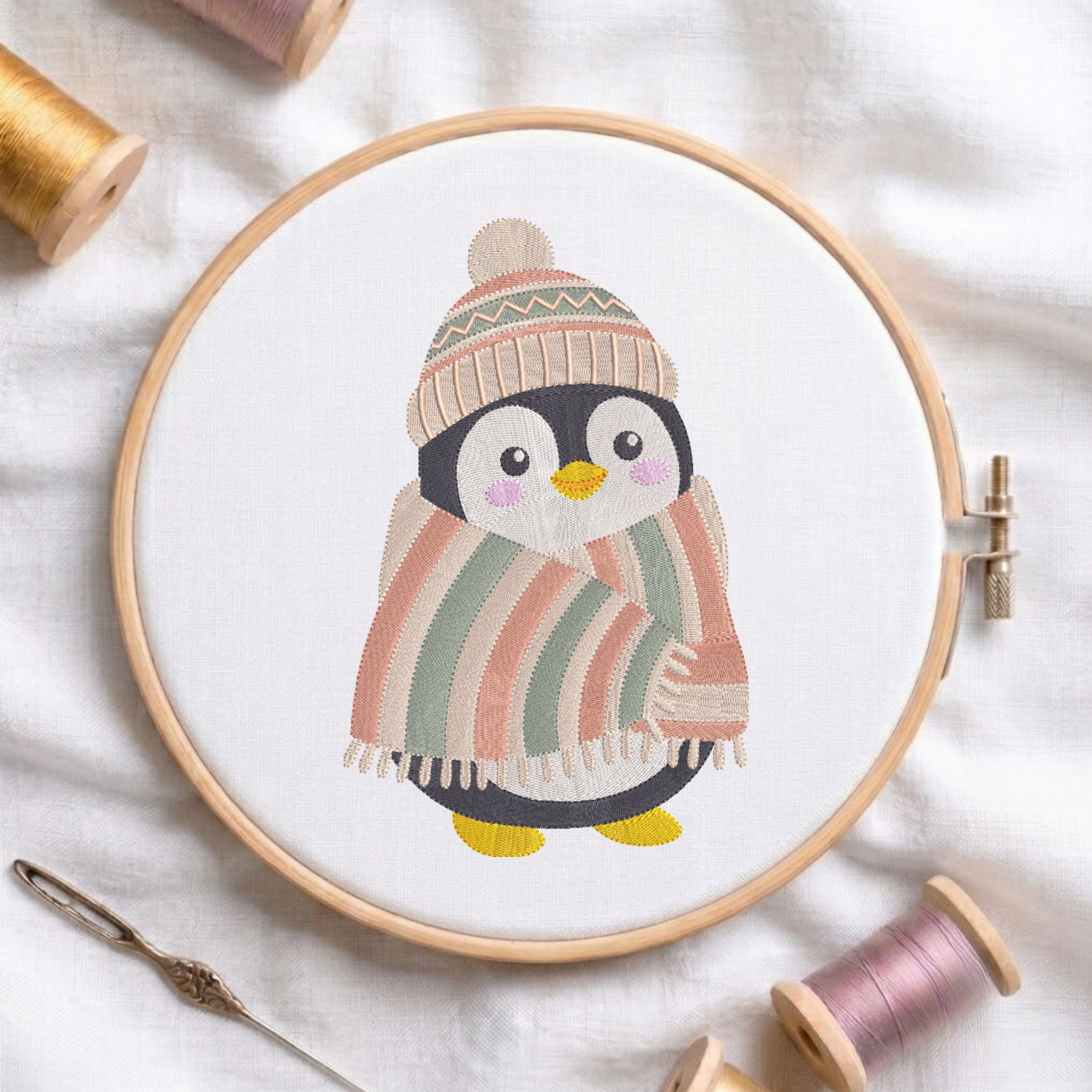A circular embroidery hoop holds a white fabric with a stitched penguin design wearing a striped knit hat and a striped blanket in soft peach, sage green, cream, black, and yellow. The hoop is photographed on white fabric with spools of thread and a needle around it.