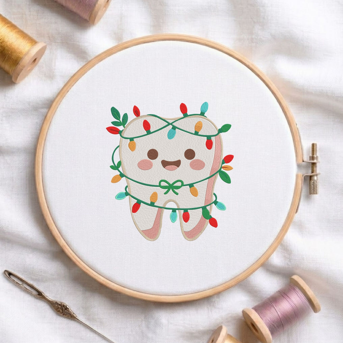 Cute smiling tooth wrapped in colorful Christmas string lights machine embroidery design shown in a wooden embroidery hoop on white fabric with thread spools and a needle