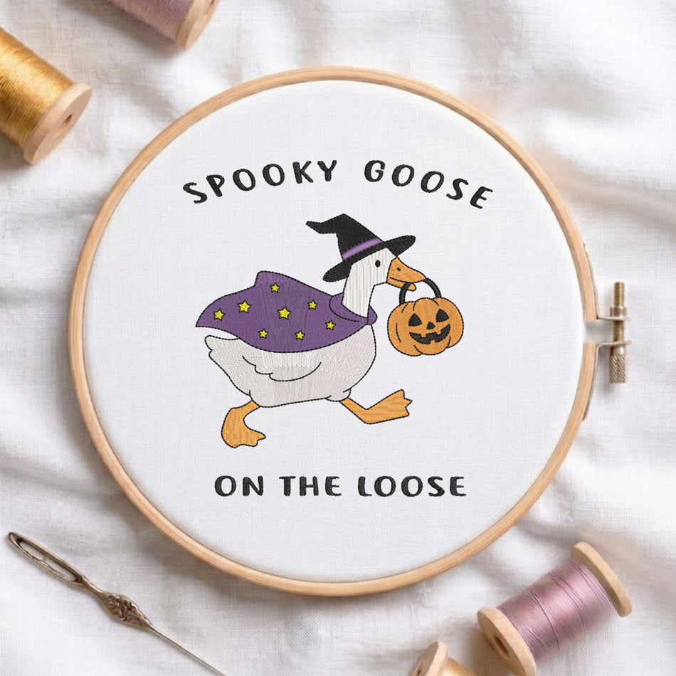 "Spooky Goose on the Loose" witch goose holding a jack-o-lantern machine embroidery design displayed in a wooden hoop on white fabric with thread spools and a needle