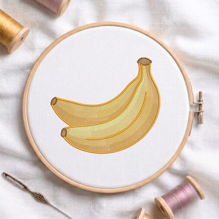 A circular embroidery hoop holding white fabric with a stitched illustration of two yellow bananas. The hoop is photographed on a white fabric surface with spools of thread and a needle around it.