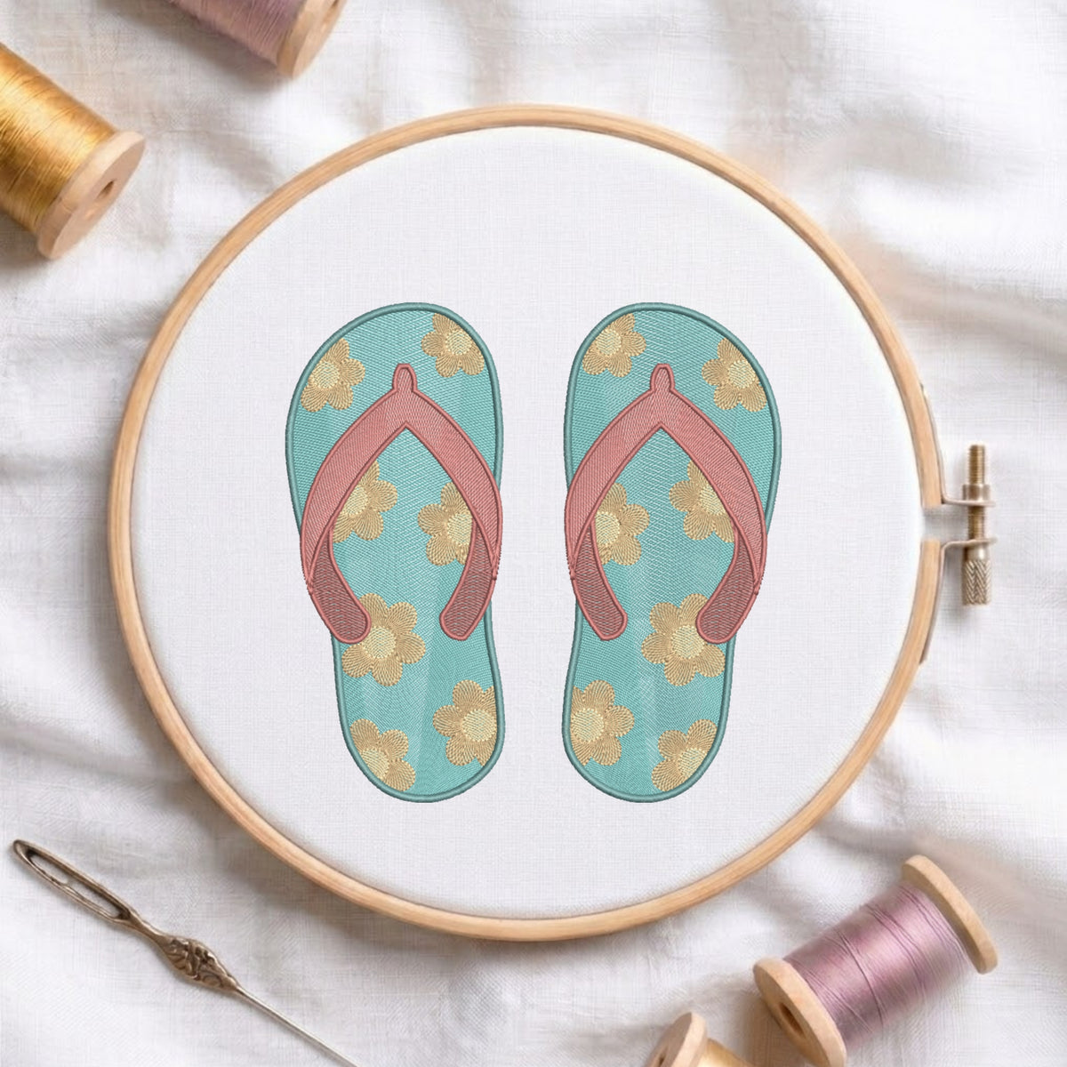 A circular embroidery hoop displays a floral flip-flop sandal design on white fabric, surrounded by spools of thread and a needle on a soft white cloth background.
