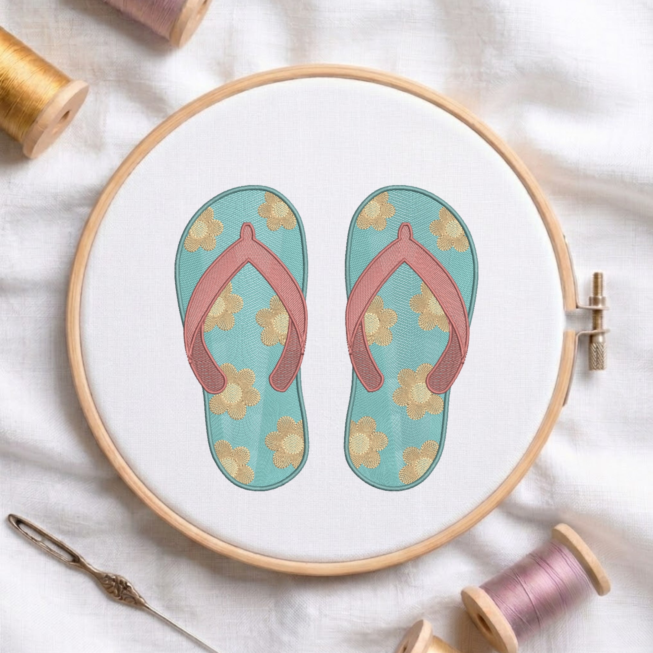 A circular embroidery hoop displays a floral flip-flop sandal design on white fabric, surrounded by spools of thread and a needle on a soft white cloth background.