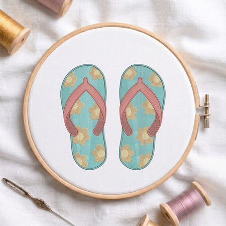 A circular embroidery hoop displays a floral flip-flop sandal design on white fabric, surrounded by spools of thread and a needle on a soft white cloth background.