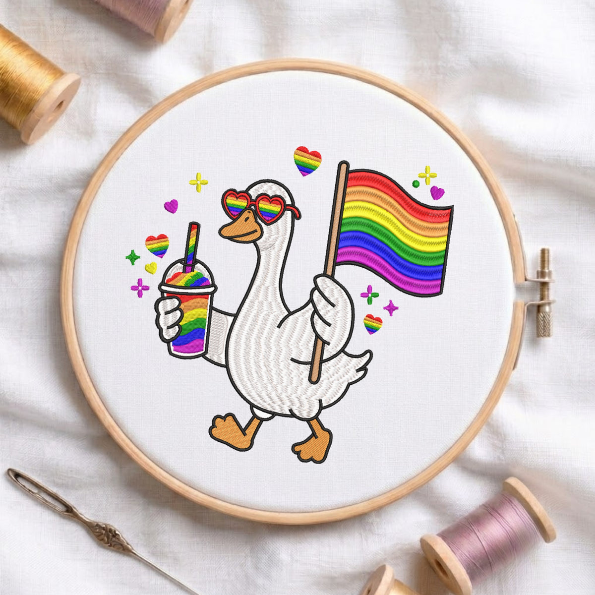 A circular embroidery hoop on white fabric, featuring a white goose wearing rainbow heart sunglasses and holding a rainbow pride flag and a rainbow drink. Spools of thread and a needle are arranged around the hoop.