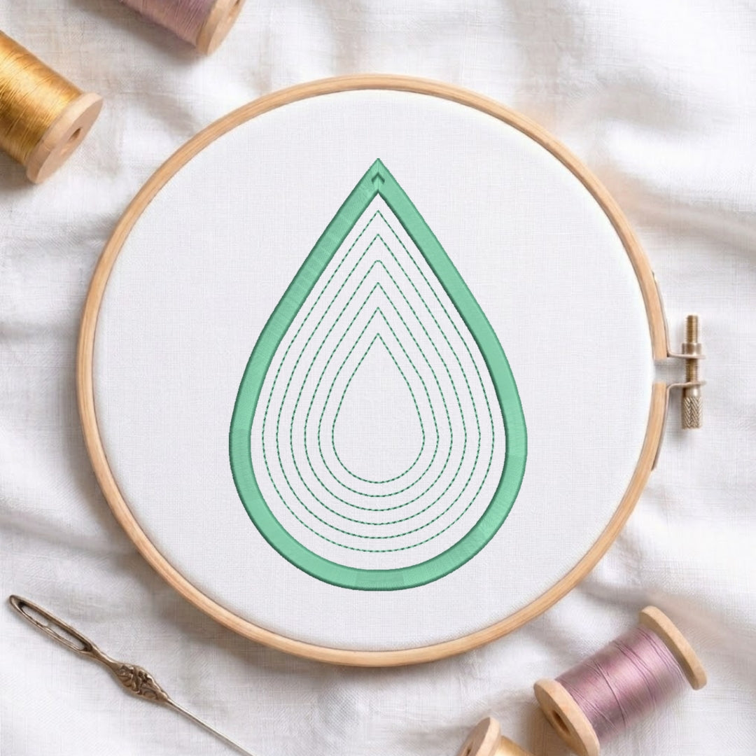 A wooden embroidery hoop holding white fabric with a mint green teardrop design outlined in stitched concentric loops. A needle lies at the lower left, and spools of thread are visible around the hoop on a white fabric background.