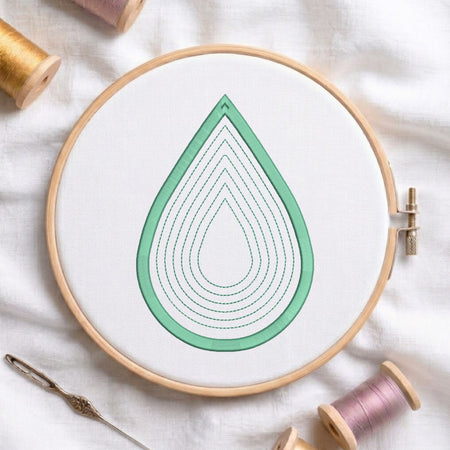 A wooden embroidery hoop holding white fabric with a mint green teardrop design outlined in stitched concentric loops. A needle lies at the lower left, and spools of thread are visible around the hoop on a white fabric background.