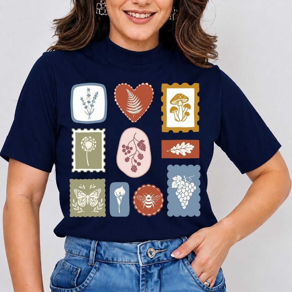 A woman wearing a navy blue t-shirt with a graphic print of nine vintage-style botanical stamps in various colors, paired with blue jeans against a light gray background.