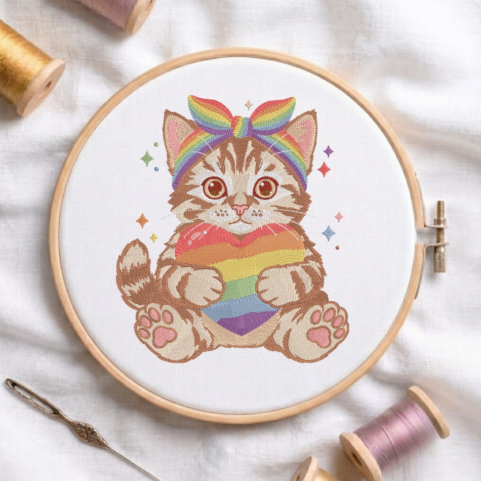 A circular embroidery hoop holds white fabric with a colorful stitched design of a striped kitten wearing a rainbow bow and holding a rainbow heart. The hoop is styled on a white fabric background with spools of thread and a needle nearby.