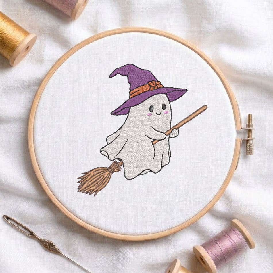 A circular embroidery hoop holding white fabric with a cute cartoon ghost wearing a purple witch hat and holding a broomstick. The hoop is styled on a white fabric background with spools of thread and a needle nearby.