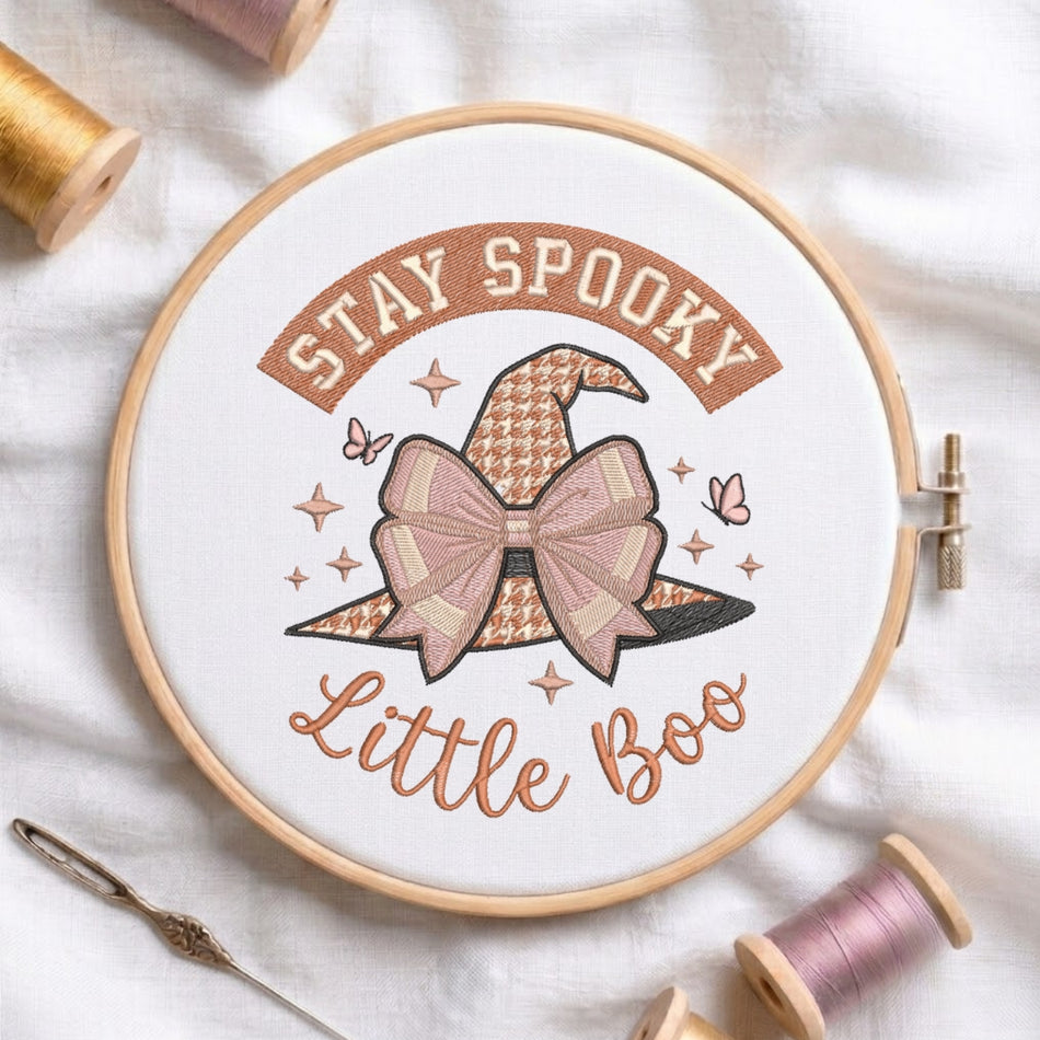 A circular embroidery hoop on white fabric, showing a stitched design with the words “STAY SPOOKY Little Boo” around a witch hat and bow. Spools of thread and a needle are arranged around the hoop.