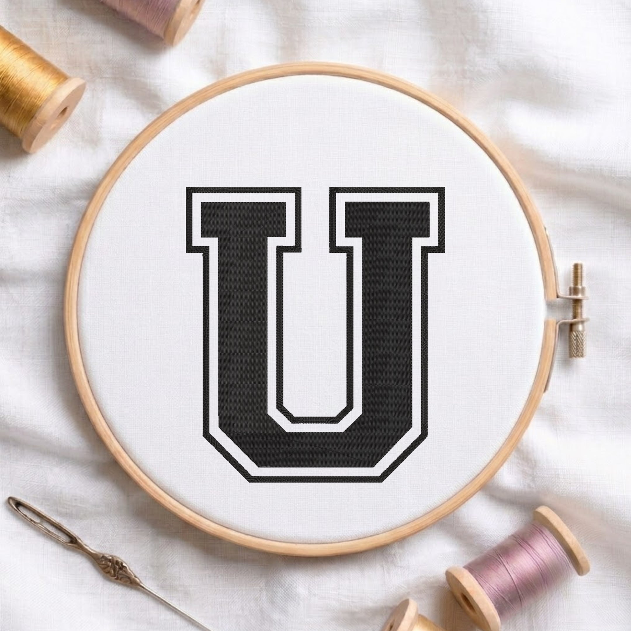 A wooden embroidery hoop holding white fabric with a large black letter U outlined in white. A needle and thread spools are arranged around it on a white fabric background.