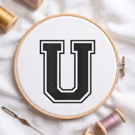 A wooden embroidery hoop holding white fabric with a large black letter U outlined in white. A needle and thread spools are arranged around it on a white fabric background.