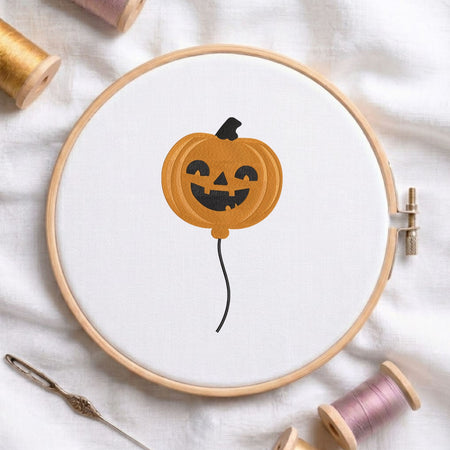 Cute ghost holding a jack-o-lantern pumpkin balloon machine embroidery design displayed in a wooden embroidery hoop on white fabric with thread spools and a needle