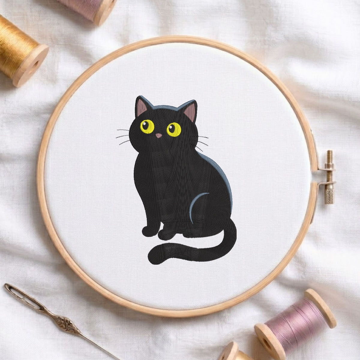 Cute black cat with big yellow eyes machine embroidery design, displayed inside a round wooden hoop on white fabric with thread spools and a needle.