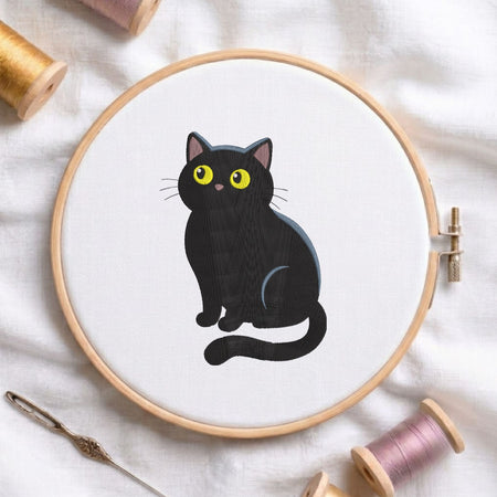 Cute black cat with big yellow eyes machine embroidery design, displayed inside a round wooden hoop on white fabric with thread spools and a needle.