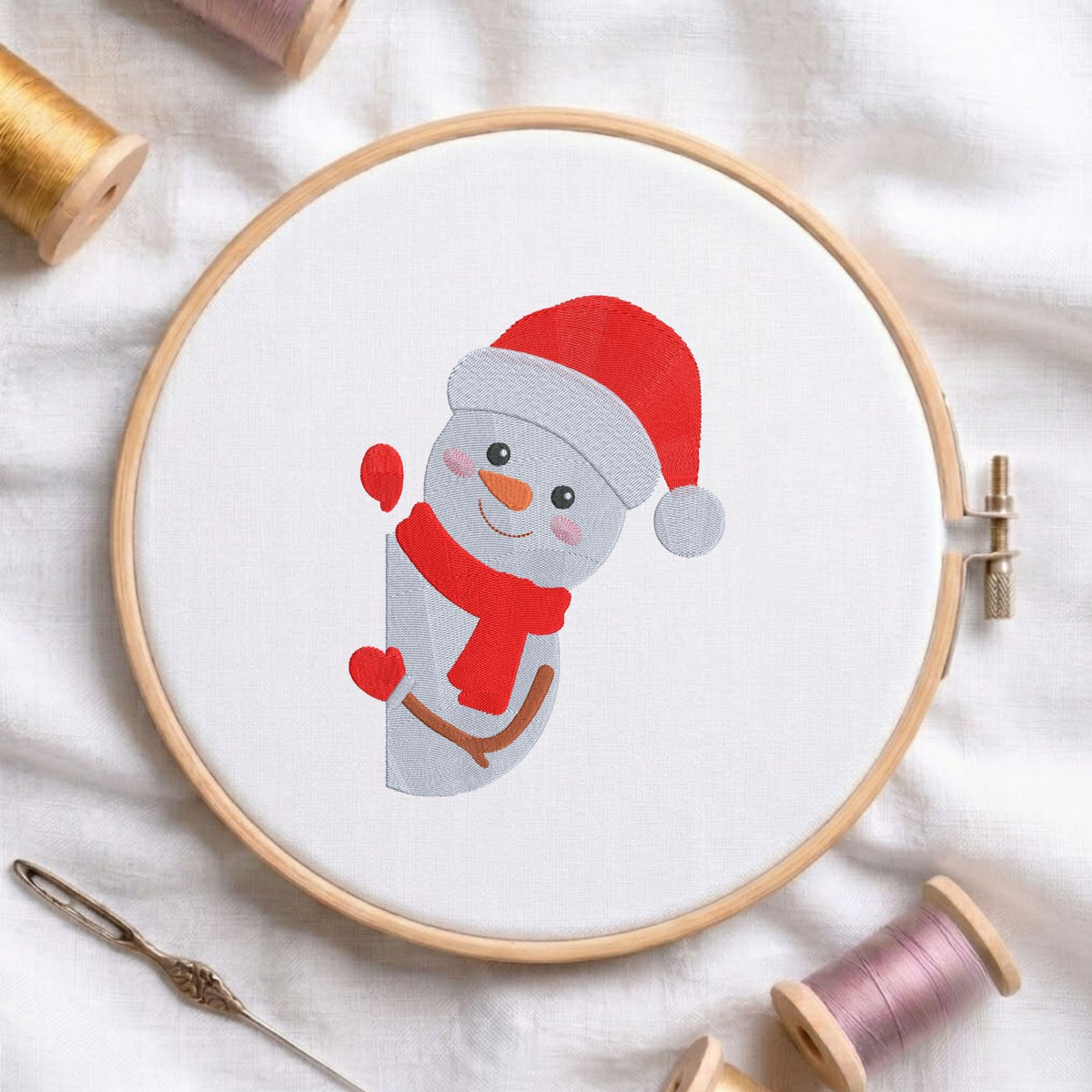 Cute snowman wearing a red Santa hat and scarf machine embroidery design displayed in a wooden hoop on white fabric with thread spools and a needle.