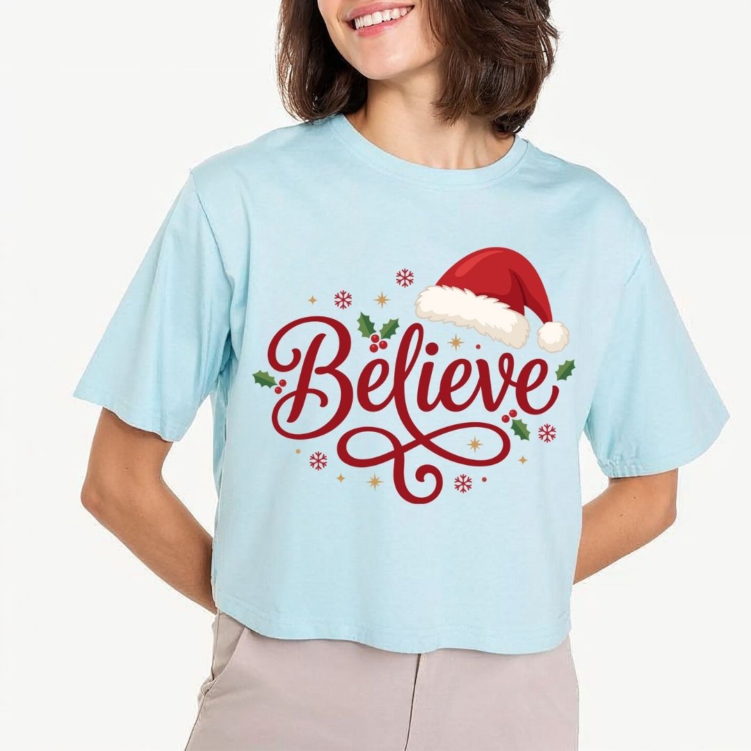 Woman wearing a light blue t-shirt featuring a red "Believe" script design with a Santa hat and holiday accents.