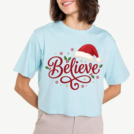 Woman wearing a light blue t-shirt featuring a red "Believe" script design with a Santa hat and holiday accents.