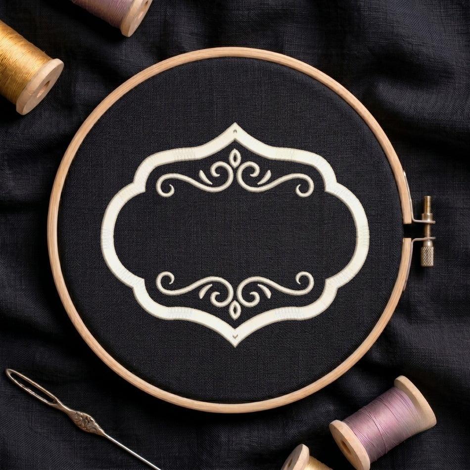 A wooden embroidery hoop holding black fabric with a white ornamental scroll design, surrounded by spools of thread and a needle on a dark fabric background.