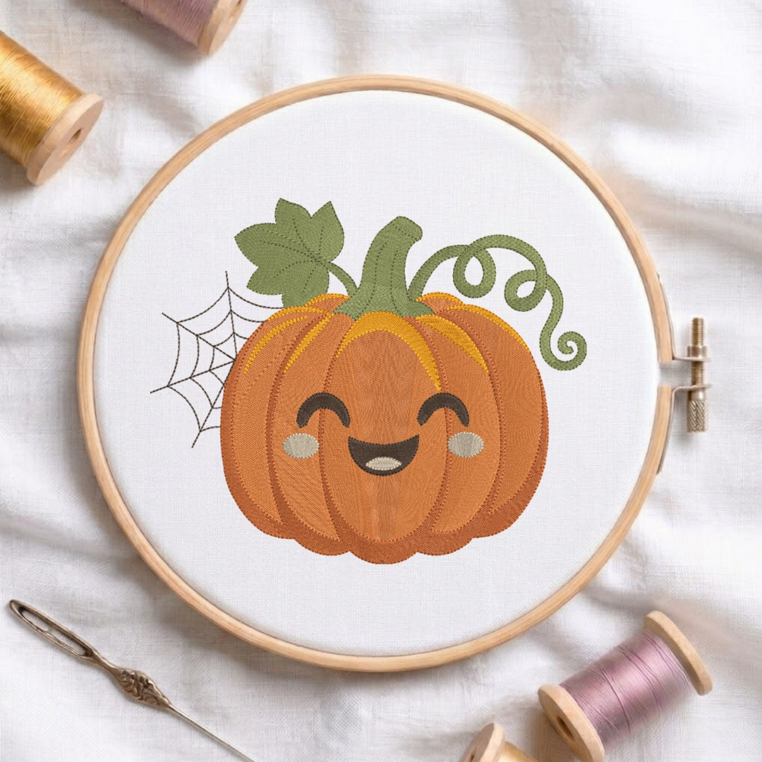 A circular embroidery hoop on white fabric displays a cute pumpkin design with a smiling face, green leaves, and a spiderweb motif. Spools of thread and a needle are arranged around it on a soft white fabric background.