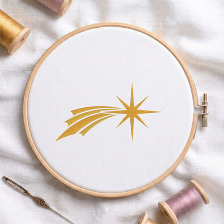 Golden comet star machine embroidery design displayed in a wooden hoop on white fabric with thread spools and a needle