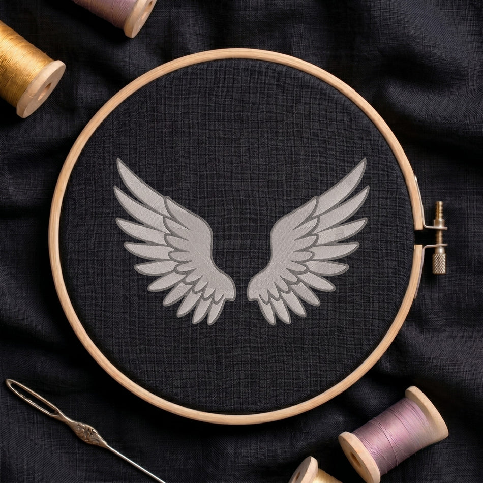 A circular embroidery hoop holding black fabric with a gray wing motif in the center, surrounded by spools of thread and a needle on a dark fabric background.