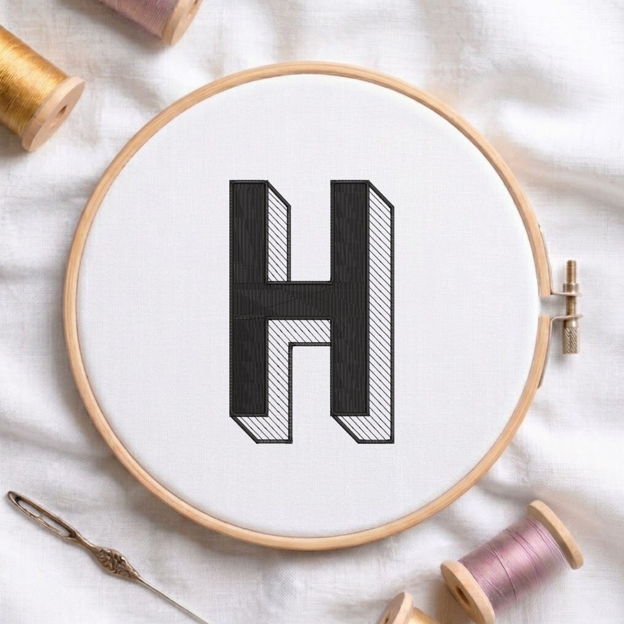 A wooden embroidery hoop holding white fabric with a large black letter H stitched in the center, surrounded by spools of thread and a needle on a white fabric background.