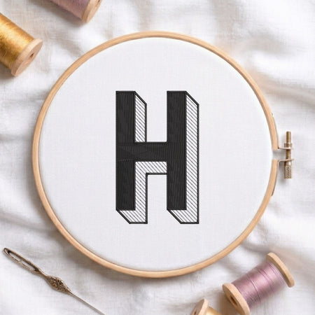 A wooden embroidery hoop holding white fabric with a large black letter H stitched in the center, surrounded by spools of thread and a needle on a white fabric background.