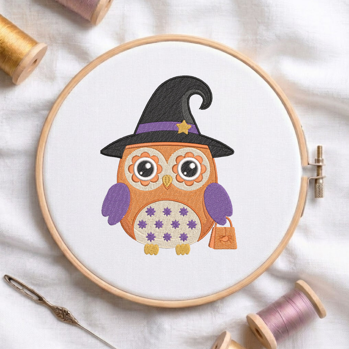 A circular embroidery hoop holding white fabric with a stitched design of an orange owl wearing a black witch hat with purple trim and a small orange bag. The hoop is styled on a white fabric background with spools of thread and a needle around it.