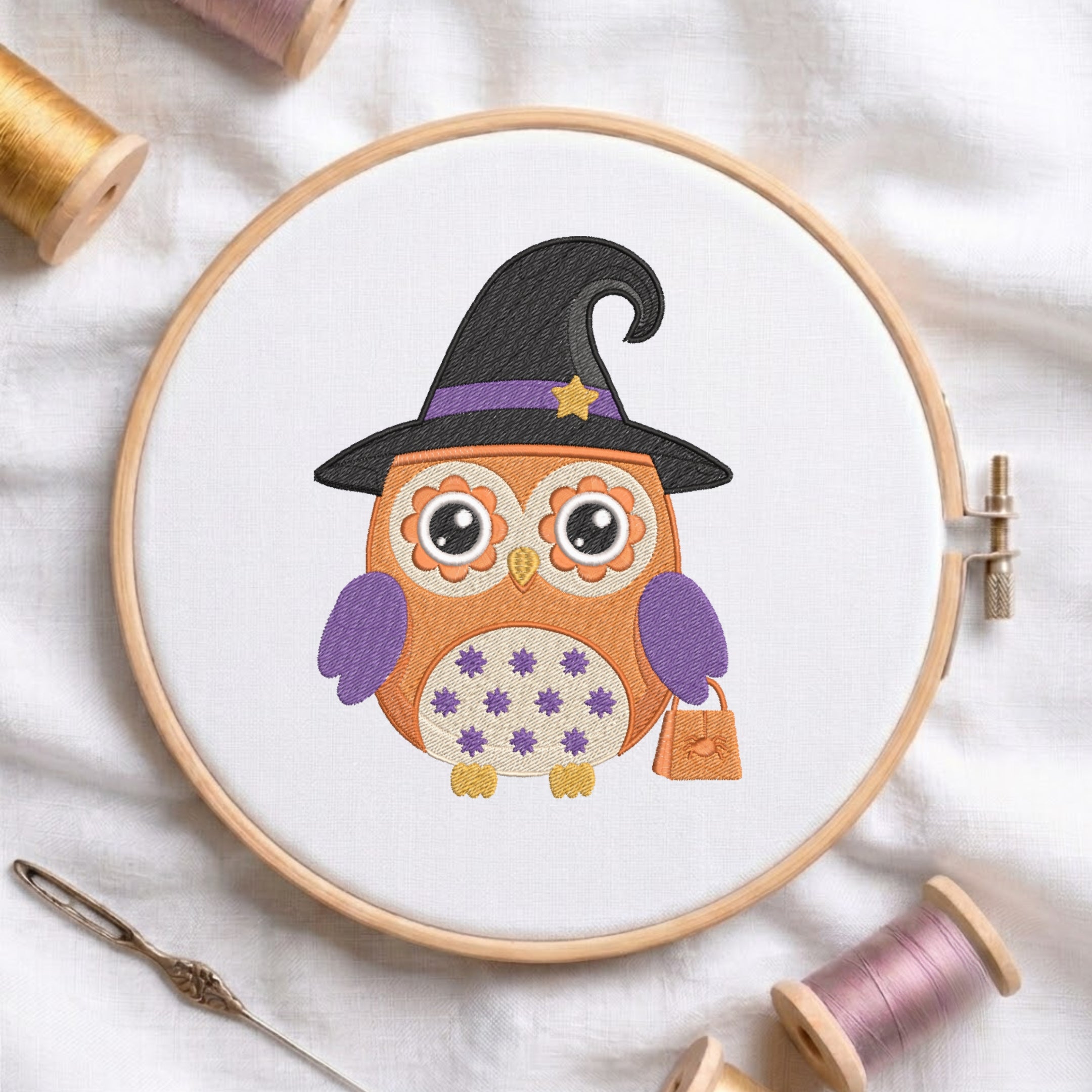 A circular embroidery hoop holding white fabric with a stitched design of an orange owl wearing a black witch hat with purple trim and a small orange bag. The hoop is styled on a white fabric background with spools of thread and a needle around it.