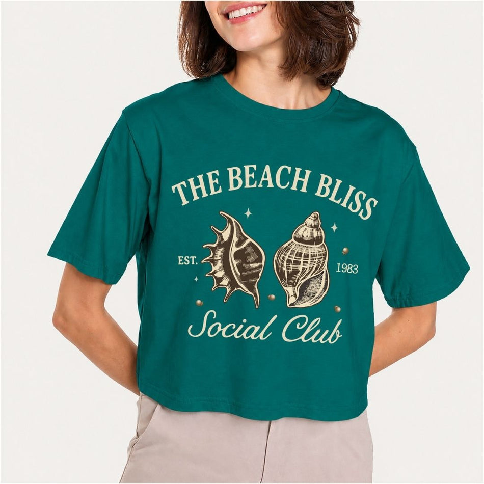 A woman smiling and wearing a dark teal green t-shirt featuring a cream-colored vintage beach club design with two detailed sketches of seashells and text reading The Beach Bliss Social Club.
