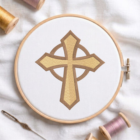 An embroidery hoop on a white cloth displays a finished machine embroidery design of a textured gold-yellow Celtic cross with an intersecting ring and a brown outline, flanked by spools of thread and a needle.