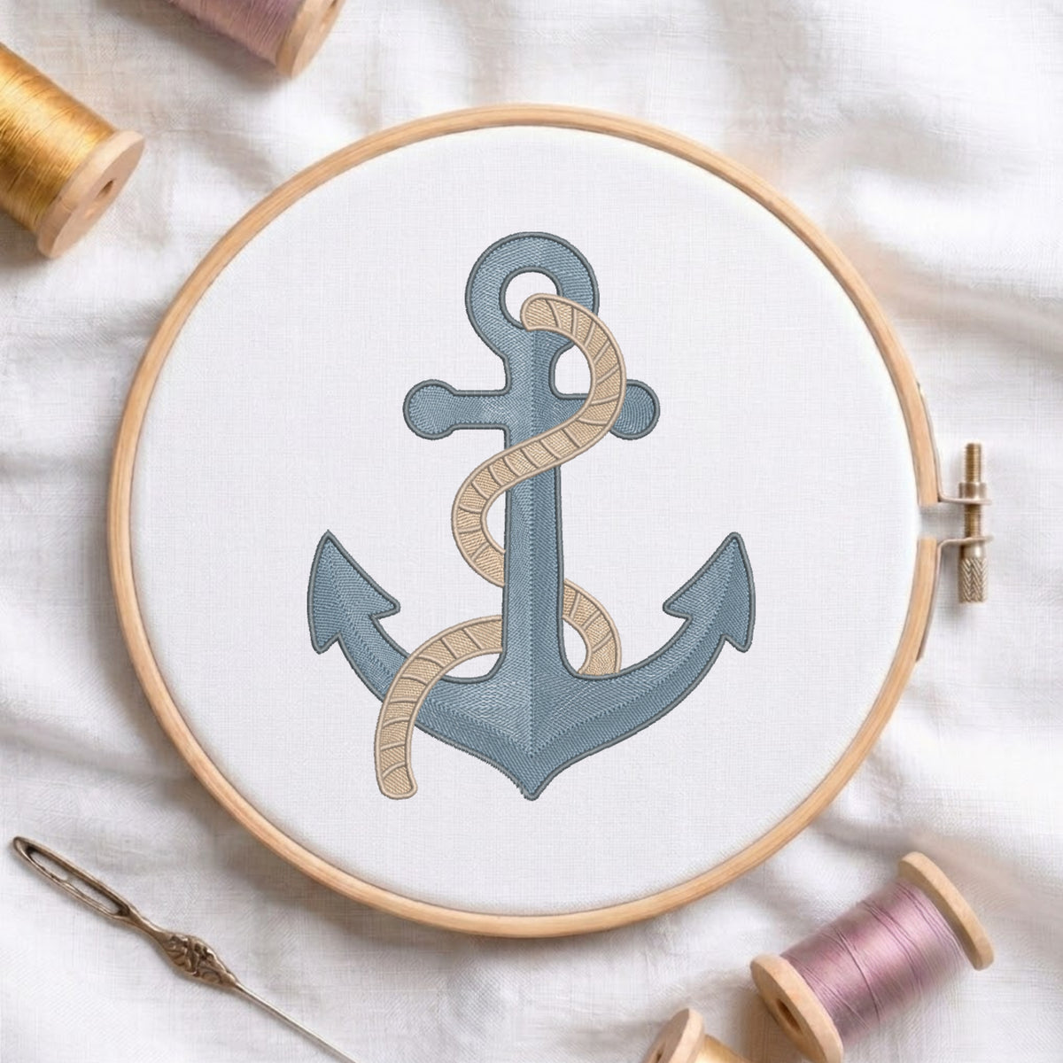 A wooden embroidery hoop holds white fabric with a stitched anchor design in muted blue-gray and beige rope, surrounded by spools of thread and a needle on a white fabric background.