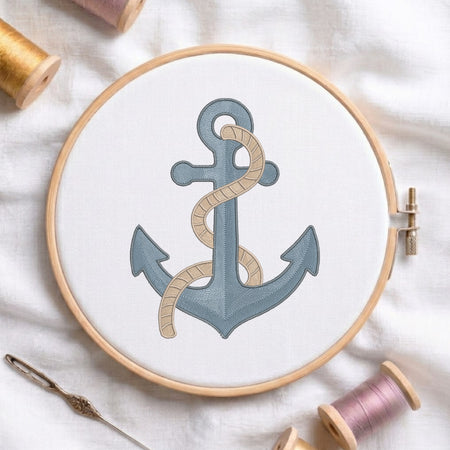 A wooden embroidery hoop holds white fabric with a stitched anchor design in muted blue-gray and beige rope, surrounded by spools of thread and a needle on a white fabric background.