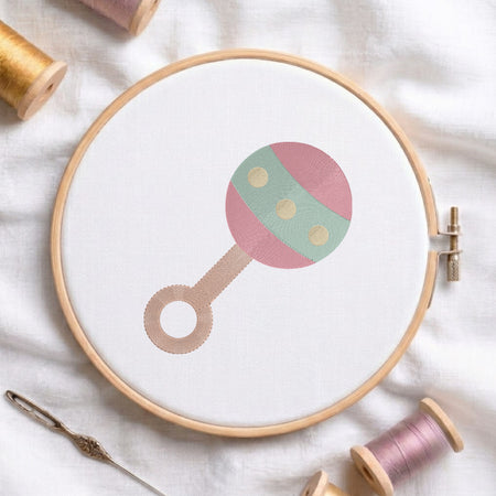 A wooden embroidery hoop holding white fabric with a stylized baby rattle design in pink, mint green, and beige. The hoop is styled on a white fabric background with spools of thread and a needle.