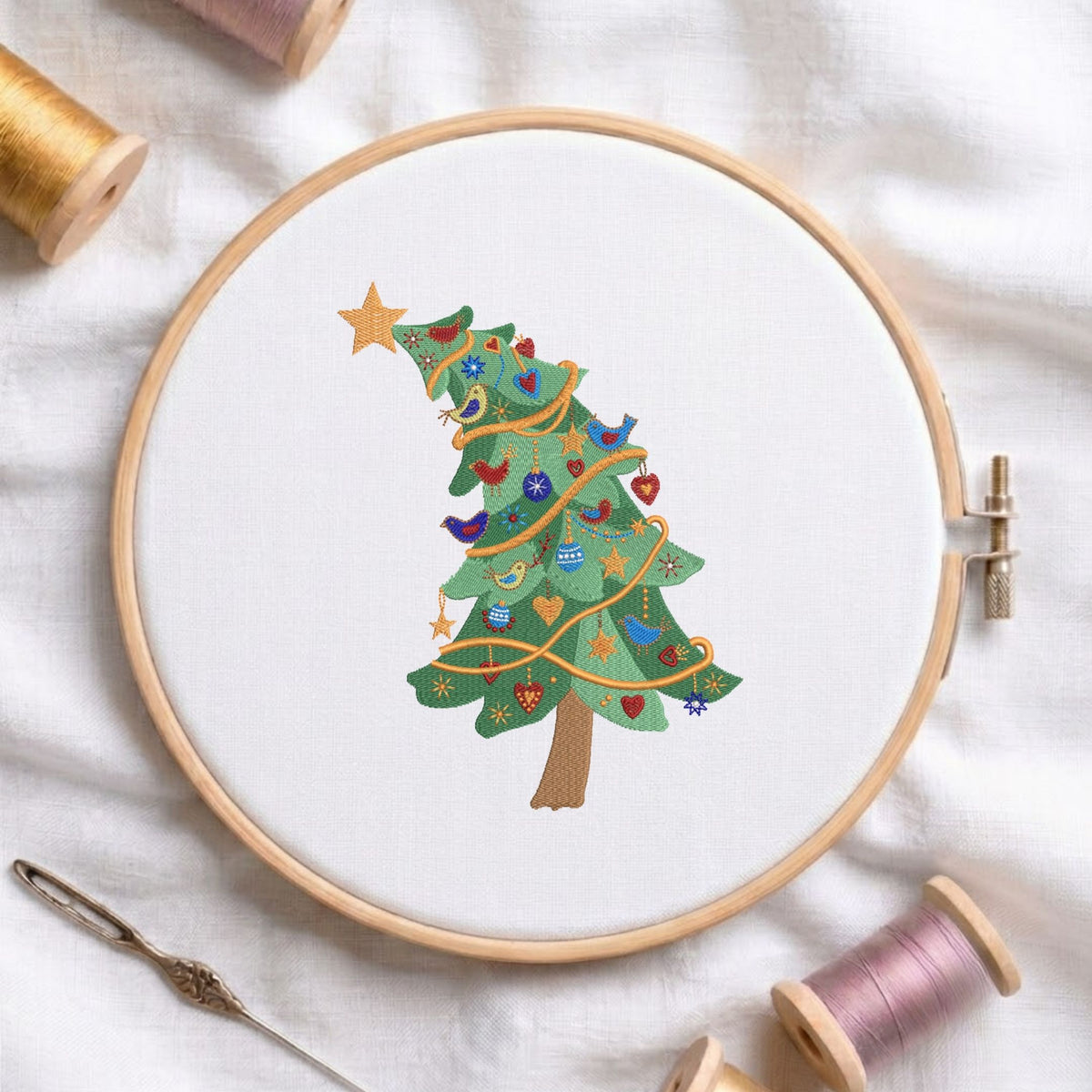 Tilted Christmas tree machine embroidery design featuring green branches wrapped in gold garland, decorated with blue birds, red hearts, and stars, displayed inside a round wooden hoop on white linen fabric alongside thread spools and a needle.