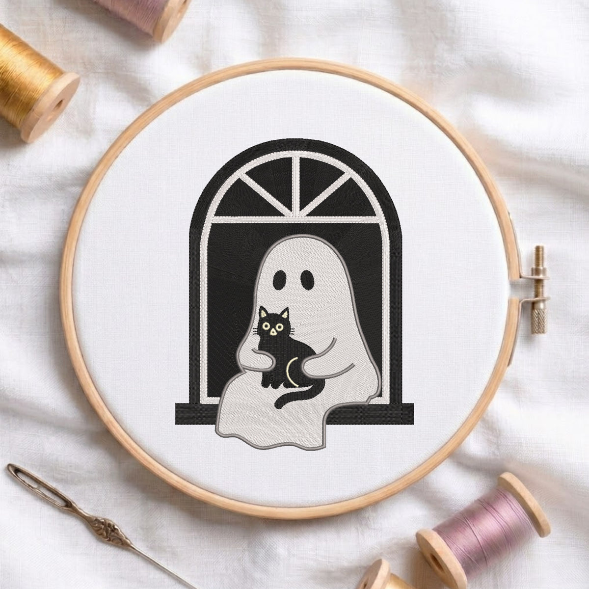 A circular embroidery hoop on white fabric, showing a black-and-white ghost holding a black cat design. Spools of thread and a needle are arranged around it.