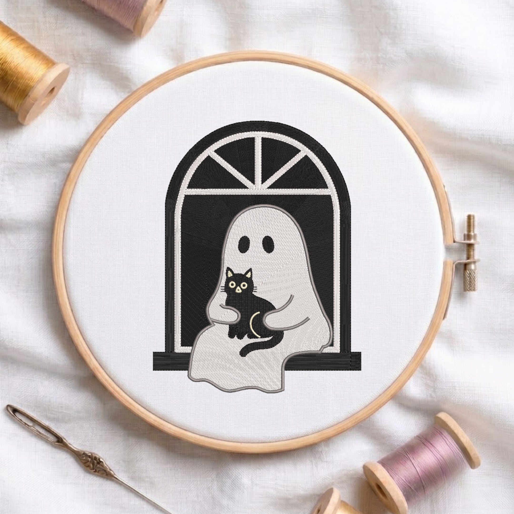 A circular embroidery hoop on white fabric, showing a black-and-white ghost holding a black cat design. Spools of thread and a needle are arranged around it.