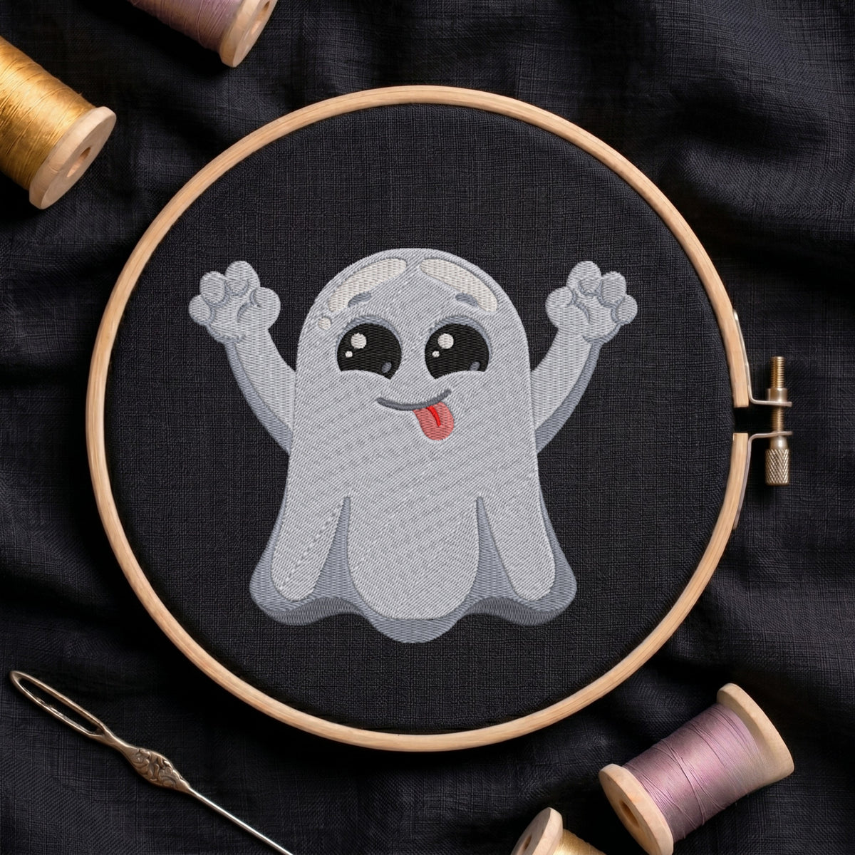 A circular embroidery hoop holding a stitched cartoon ghost design on black fabric, surrounded by spools of thread and a needle on a dark cloth background.