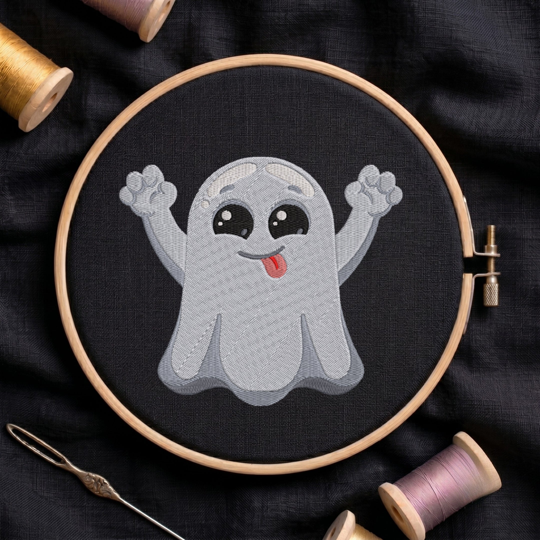 A circular embroidery hoop holding a stitched cartoon ghost design on black fabric, surrounded by spools of thread and a needle on a dark cloth background.