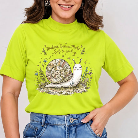 Smiling person wearing a white t-shirt featuring a cute illustrated snail with a patterned shell and the text "Makers Gonna Make... S-l-o-w-l-y" surrounded by botanical accents.