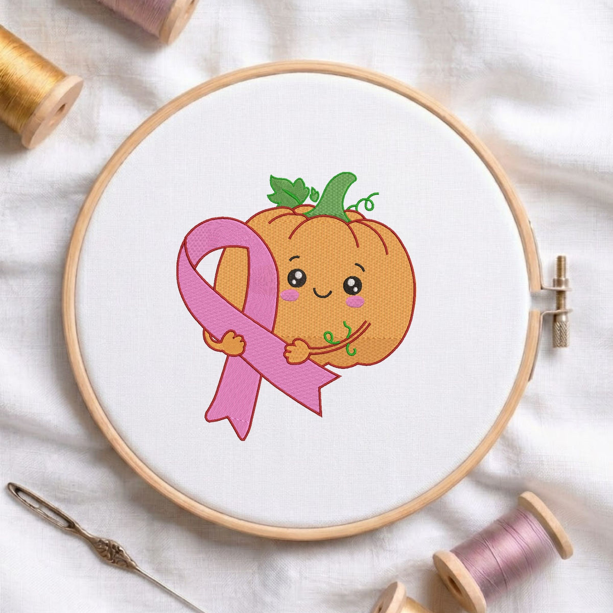 Cute kawaii pumpkin character hugging a pink breast cancer awareness ribbon machine embroidery design, displayed in a wooden hoop on white fabric with thread spools and a needle.