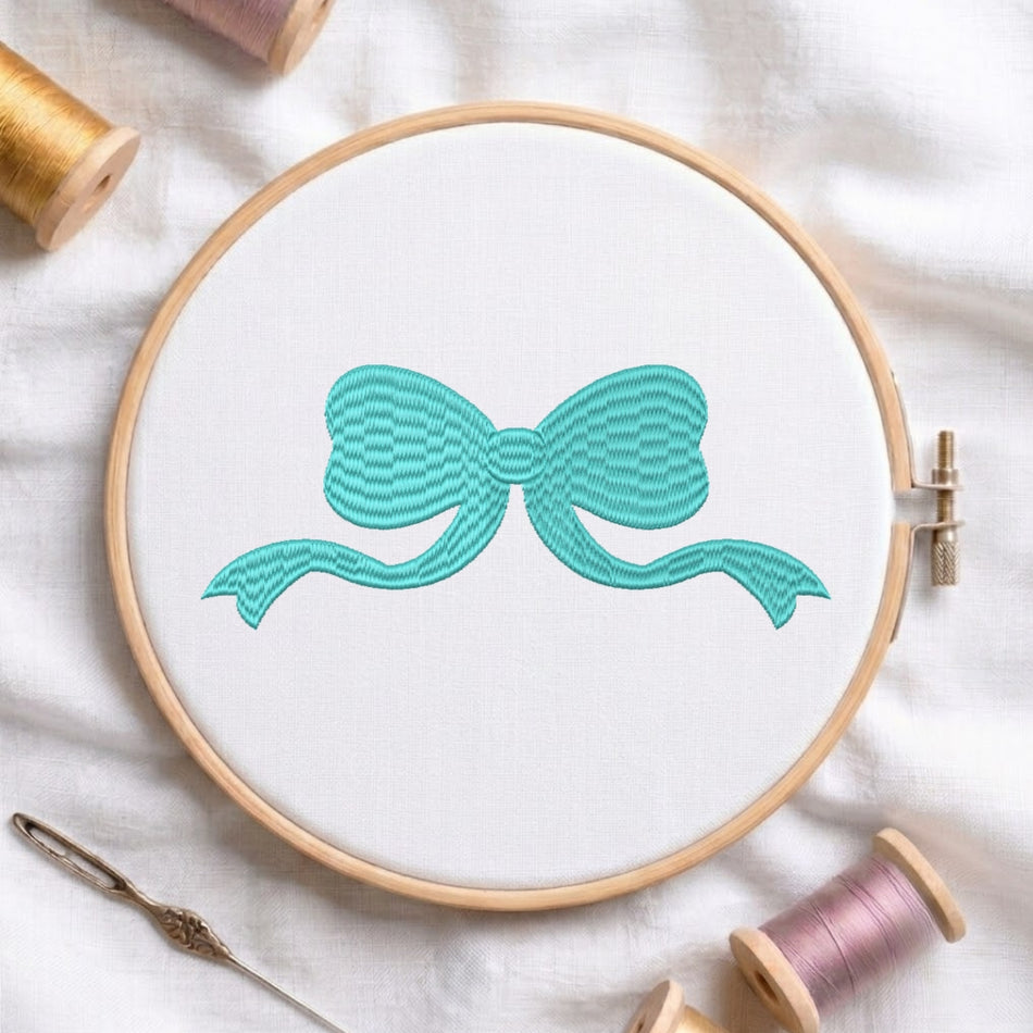 A wooden embroidery hoop holding white fabric with a turquoise bow design, surrounded by spools of thread and a needle on a white fabric background.
