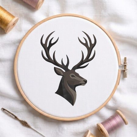 A circular embroidery hoop holding white fabric with a stylized deer head design in dark gray and brown. The hoop is styled on a white fabric background with spools of thread and a needle nearby.