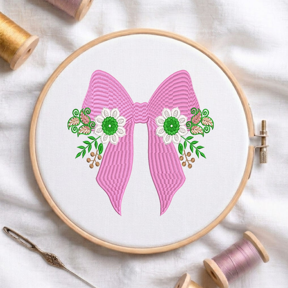 A circular embroidery hoop holding white fabric with a pink bow design, decorated with green leaves, white flowers, and small brown birds. Spools of thread and a needle are arranged around it on a white fabric background.