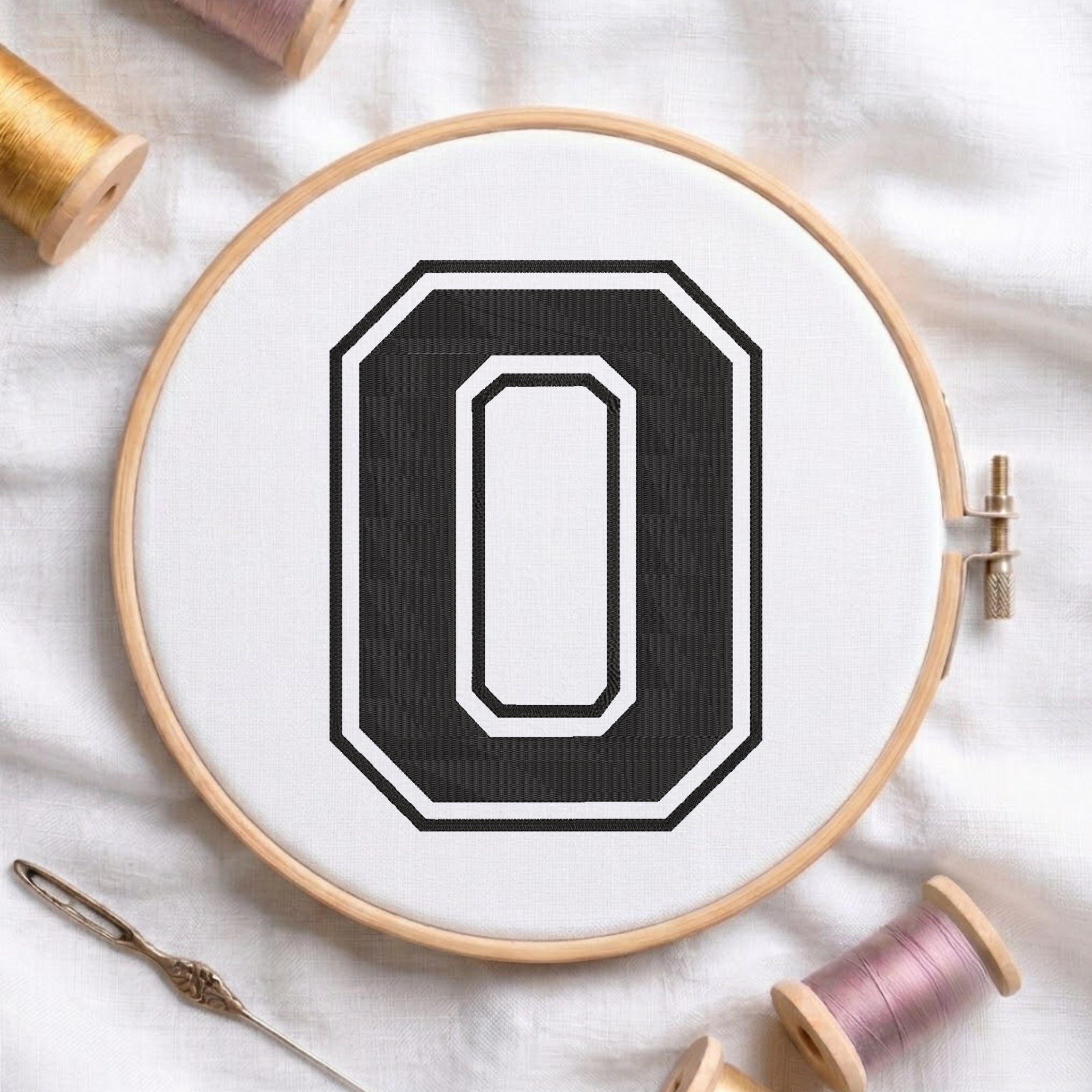 A wooden embroidery hoop holding white fabric with a large black letter O design, styled on a white cloth with spools of thread and a needle around it.
