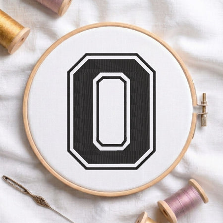 A wooden embroidery hoop holding white fabric with a large black letter O design, styled on a white cloth with spools of thread and a needle around it.